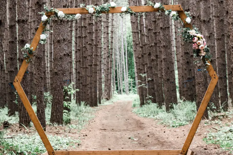 A hexagon wooden wedding arch with some flowers and greenery in the woods.